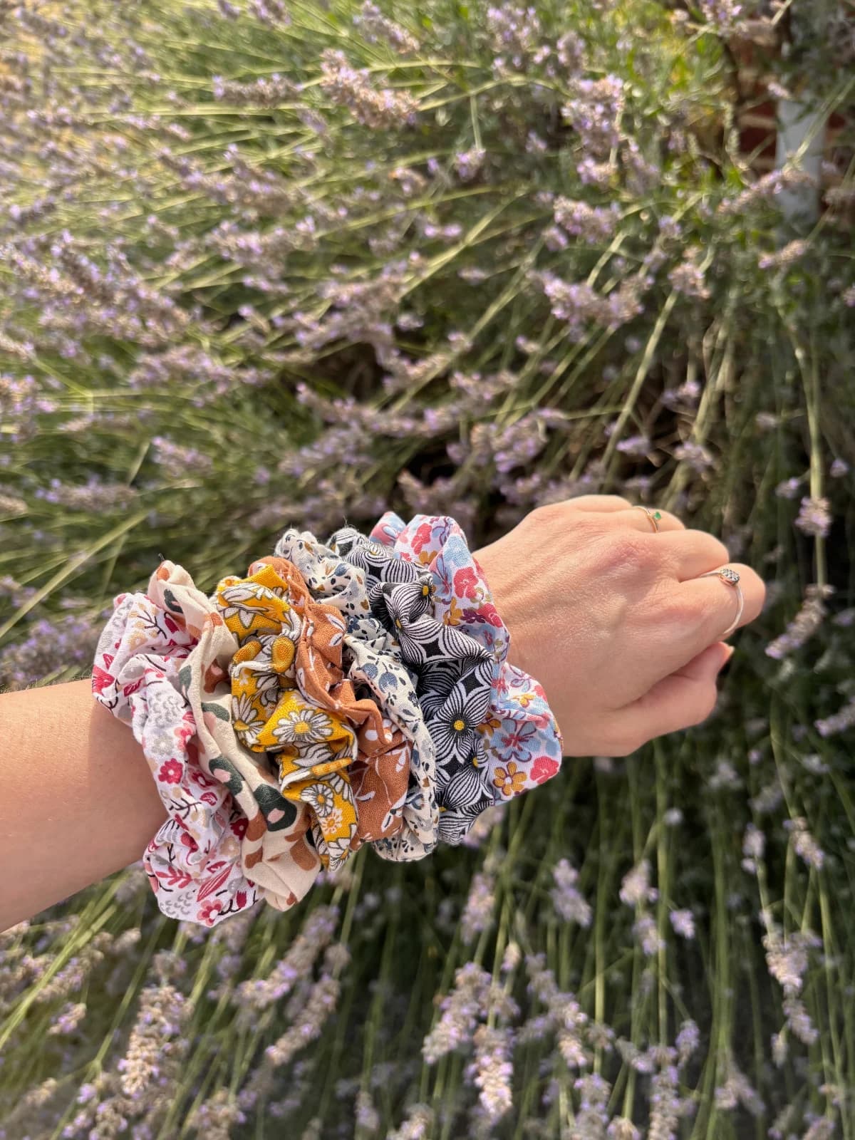 Poignet orné de plusieurs chouchous en tissu aux motifs floraux et colorés, devant un champ de lavande.