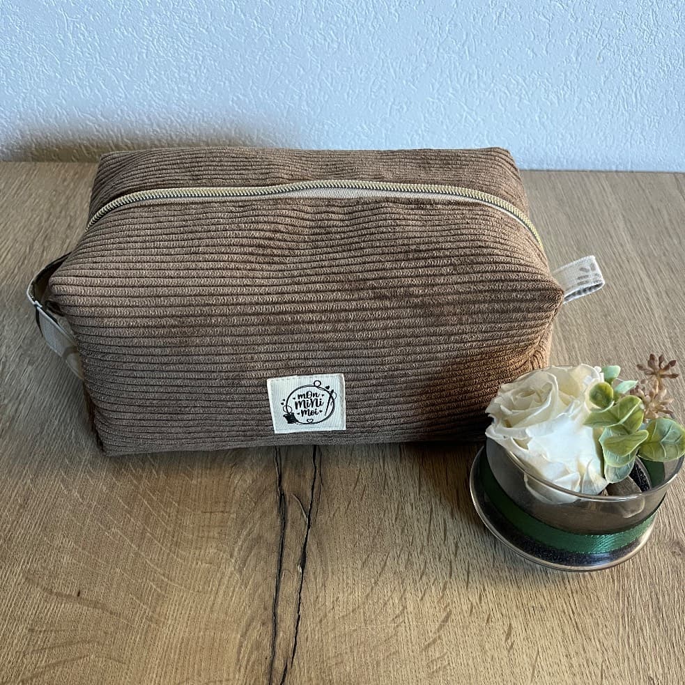 Trousse de toilette rectangulaire en velours côtelé marron fermée, posée sur une table en bois.