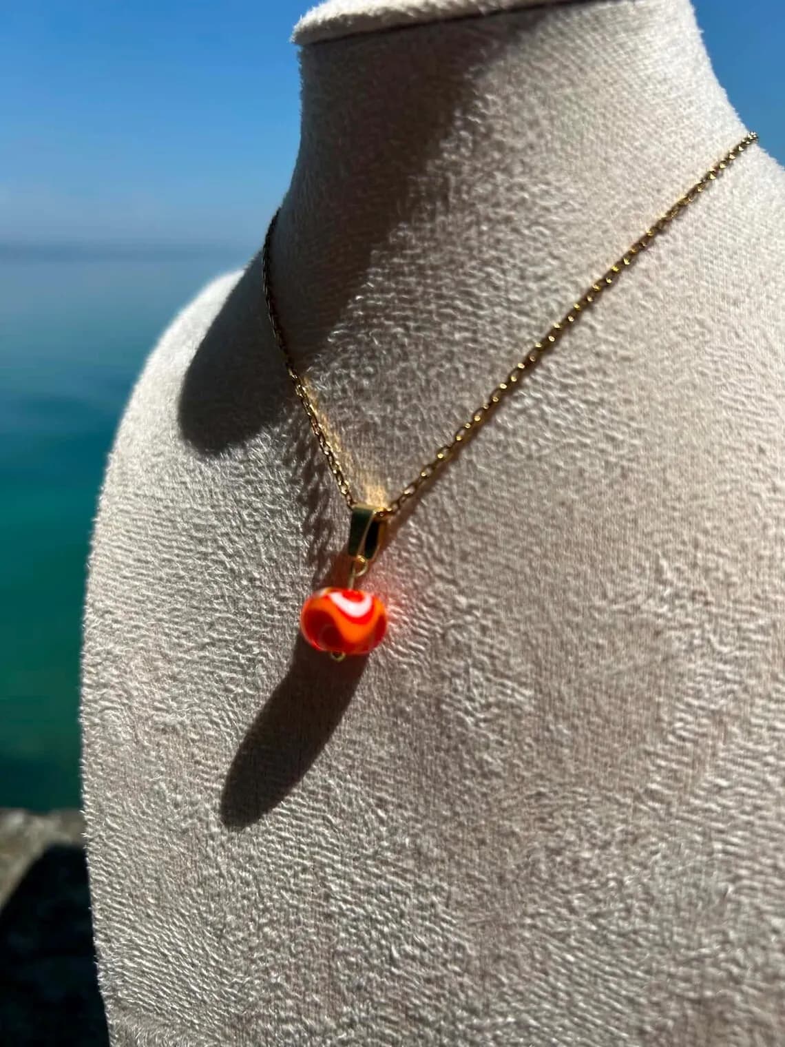 Collier avec perle en verre orange et motifs blancs sur chaîne dorée présenté sur buste face à la mer.