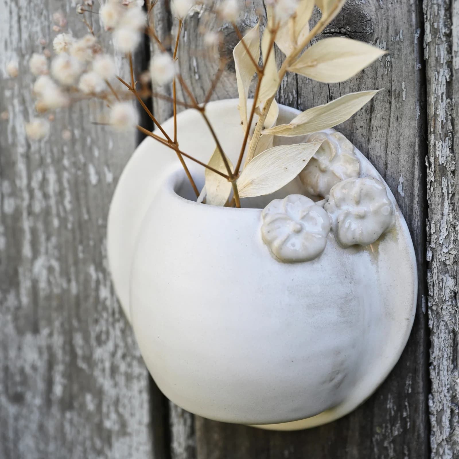 Vase mural rond en céramique blanche orné de fleurs en relief, fixé sur du bois avec des fleurs séchées.