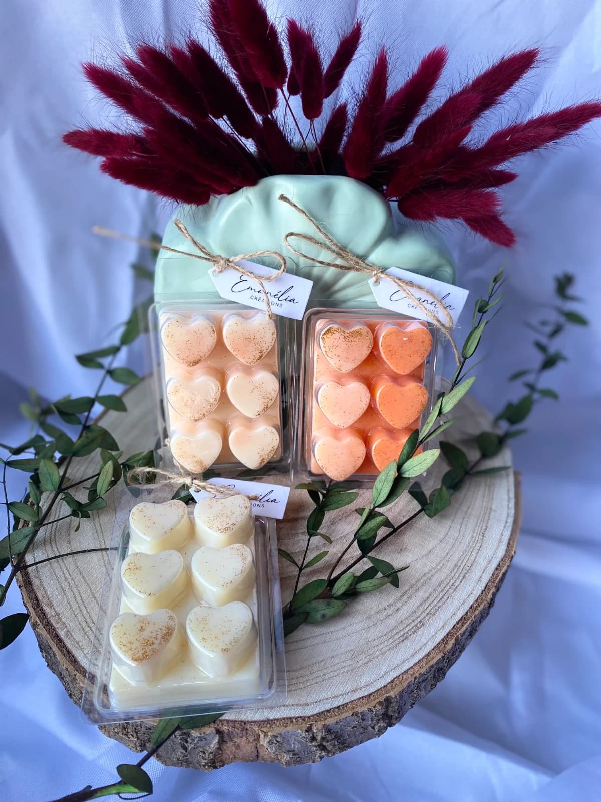 Trois boîtes clamshells de cire en forme de cœurs pailletés crème et orange sur un rondin de bois décoré.